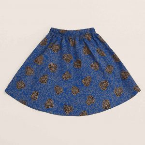 A young African girl wearing a bold blue Kitenge skirt with golden patterns, standing confidently in a studio setting with a natural smile.