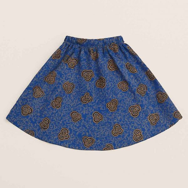 A young African girl wearing a bold blue Kitenge skirt with golden patterns, standing confidently in a studio setting with a natural smile.