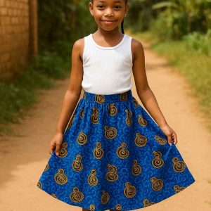 Young African girl wearing a bold Kitenge skirt in a natural outdoor setting, smiling softly and standing near a garden path.