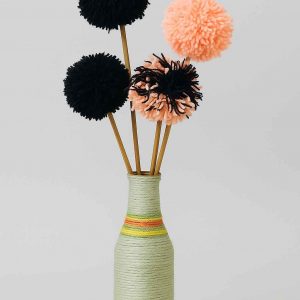 Studio image of a decorative yarn-wrapped bottle with a full arrangement of handcrafted pompom yarn flowers